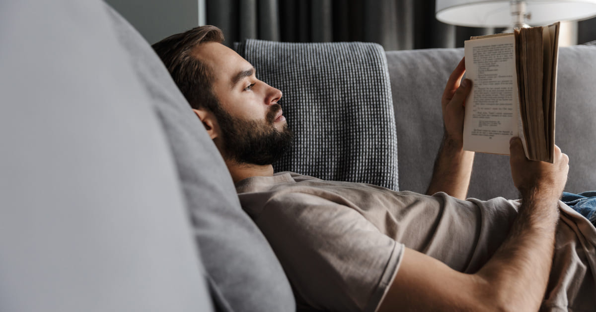 I generi di libri più letti. Uomo con la barba sdraiato su un divano legge un libro alla luce di una lampada