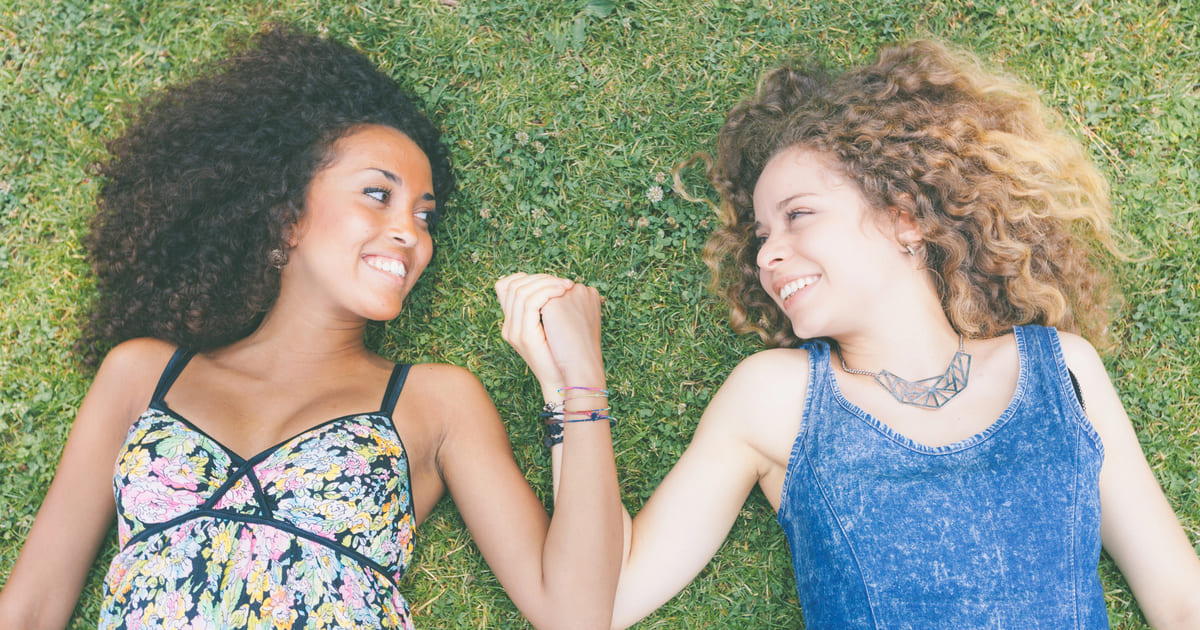 Due ragazze sdraiate a terra su un prato si tengono per mano e si guardano sorridendosi: è una interpretazione perfetta per i libri d'amore lesbico