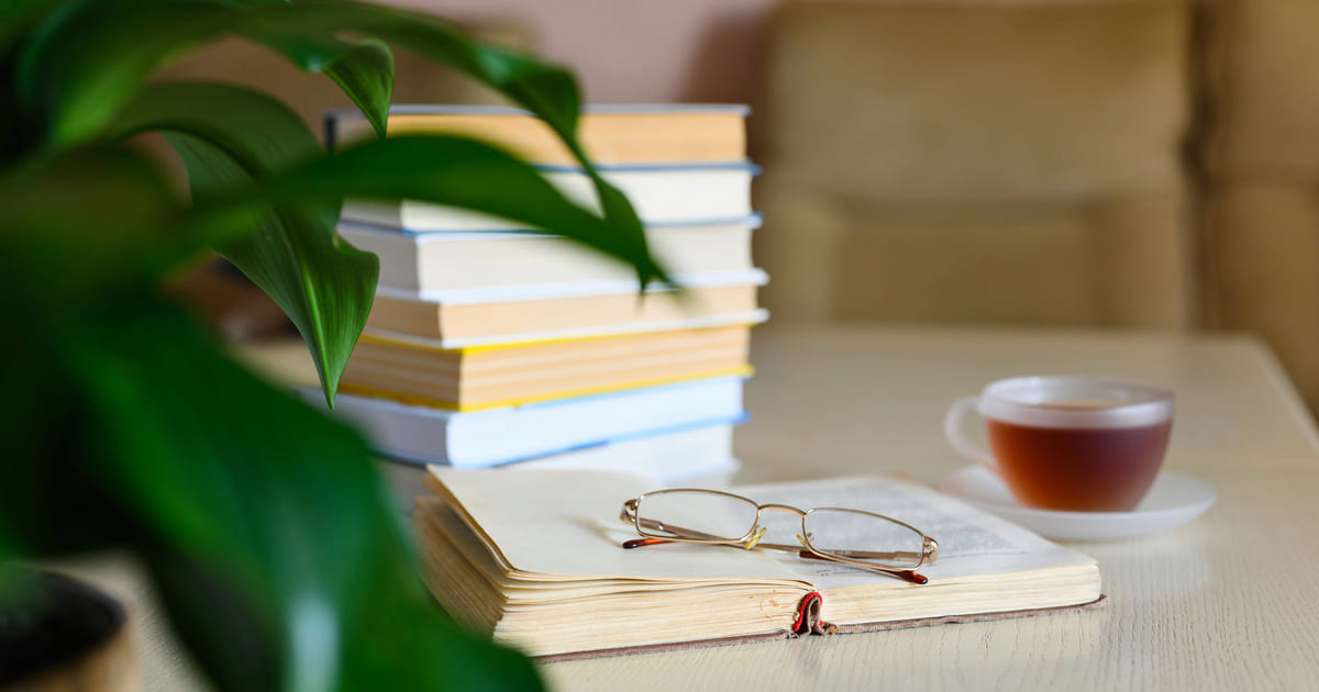 Pile di libri e occhiali su un tavolo con una tazza di tè e una pianta, simbolo di lettura e relax per scoprire le nostre mini recensioni di libri